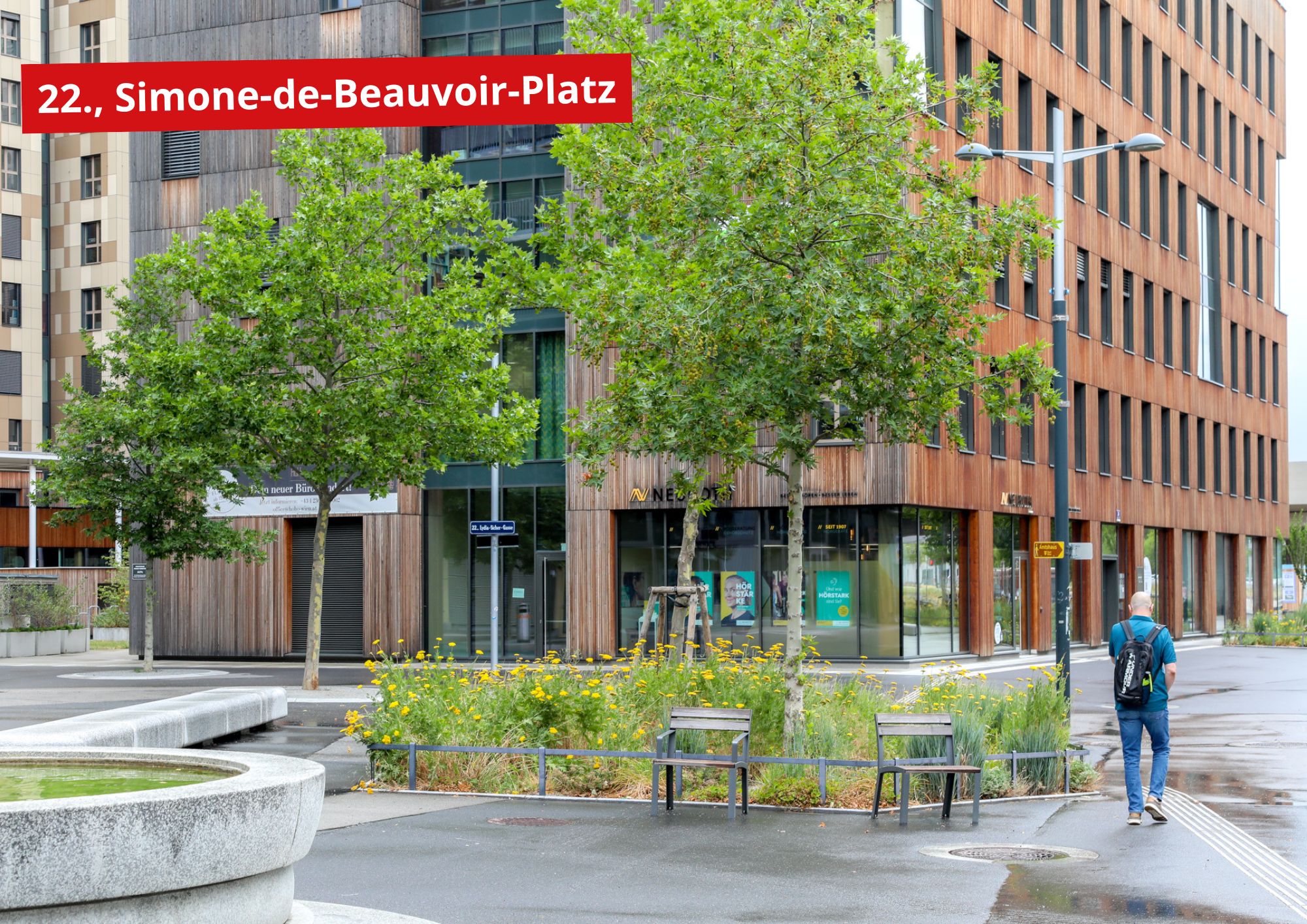 Simone-de-Beauvoir-Platz 1 Ein Mann, nur von hinten sichtbar, geht über den Simone-de-Beauvoir-Platz, auf dem schattige Bäume, ein begrüntes Blumenbeet, Sitzgelegenheiten sowie der Teil eines Brunnens zu sehen sind.